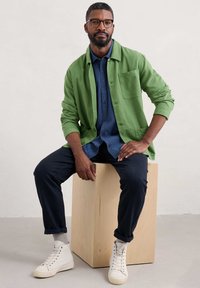 Jachetă verde cu nasturi deasupra unei cămăși texturate albastre, asociată cu pantaloni bleumarin și adidași albi cu talpă înaltă, așezată pe un cub din lemn.