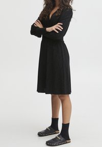 Czarna, długosleeve sukienka z głębokim dekoltem w kształcie V, teksturowana tkanina i odcięta talia. Połączona z czarnymi skarpetkami i klapkami slip-on z złotymi akcentami.
