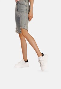 Pantaloni bermuda di jeans con orlo sfrangiato, abbinati a sneaker bianche con dettagli neri e occhielli metallici.
