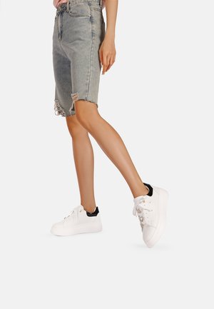 Pantaloni bermuda di jeans con orlo sfrangiato, abbinati a sneaker bianche con dettagli neri e occhielli metallici.