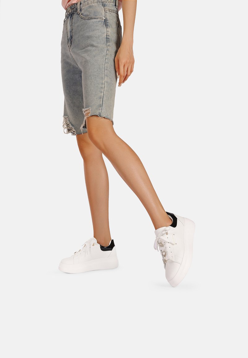 Pantaloni bermuda di jeans con orlo sfrangiato, abbinati a sneaker bianche con dettagli neri e occhielli metallici.