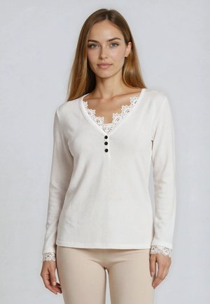 Frau mit langen Haaren, die ein weißes Langarmshirt mit Spitzenbesatz am Ausschnitt und an den Bündchen trägt und vor einem einfarbigen Hintergrund posiert.