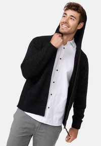 Schwarzes Strickhoodie mit Reißverschluss, gerippten Bündchen und glatter Textur. Über einem weißen Hemd mit Knopf getragen und kombiniert mit hellgrauten Hosen.