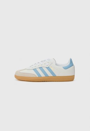 White sneakers with blue stripes, suede toe cap, and gum sole. Features laces, soft textile upper, and prominent branding on the side.