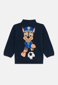 Marineblauwe fleece pullover met een grote geborduurde afbeelding van een hond in een blauwe uniform, die een zwart-wit voetbal schopt.