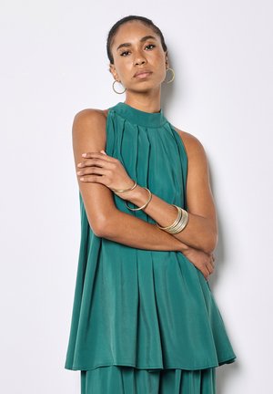 Femme aux cheveux tirés en arrière, portant un haut sans manches bleu sarcelle plissé et un pantalon large assorti, accessoirisés avec des boucles d'oreilles créoles en or et des bracelets.