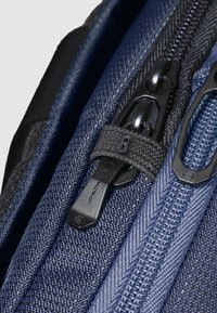 Mochila de tela azul marino con dos cremalleras negras. Superficie texturizada con patrones tejidos y un tirador de cremallera decorativo; asegurada con una correa de tela.