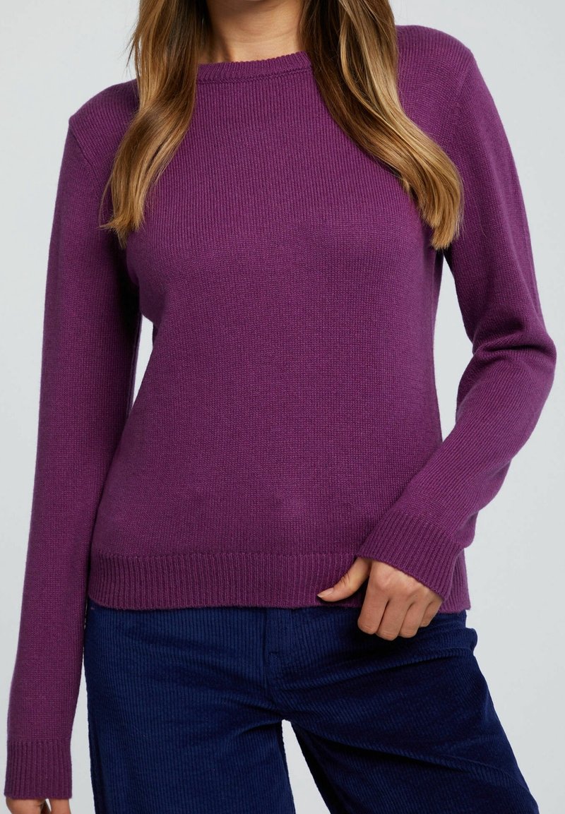 Femme portant un pull en maille violet à manches longues et un pantalon en velours côtelé bleu foncé, sa main droite touchant l'ourlet du pull.