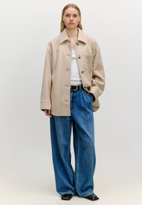 Beige Wollmischung Hemdjacke mit Knopfleiste und einer Brusttasche, kombiniert mit lockeren blauen Denim-Jeans und schwarzen Flachschuhen.