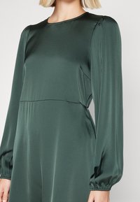 Pitkähihaiset haalari tummanvihreää satiinia. Ominaisuuksia ovat pyöreä kaula-aukko, kerätyt hihat ja rento istuvuus vartalossa. Silkkinen pinta.