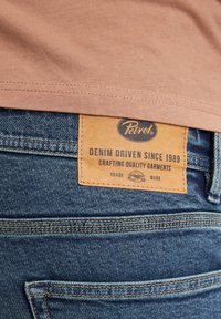 Jeans in denim con etichetta in pelle cucita sul retro, caratterizzati da tessuto blu, cuciture arancioni e il testo "Petrol" e "DENIM DRIVEN SINCE 1989."