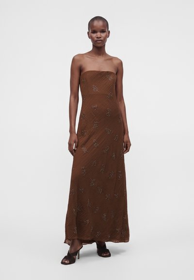 Femme debout portant une robe longue marron bustier avec une broderie florale subtile et des sandales noires à talons ouverts sur un fond uni.