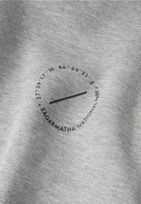 Camiseta de tela gris con un gráfico negro circular que muestra coordenadas geográficas y el texto "Parque Nacional Sagarmatha". Textura suave.