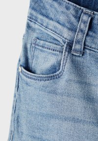 Los jeans de mezclilla azul claro presentan un bolsillo frontal, pasadores para cinturón y costuras visibles. La textura es suave con un acabado ligeramente desgastado.