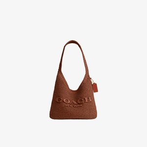 Sac à bandoulière tressé marron Coach avec une large bandoulière et une petite étiquette suspendue sur fond blanc uni.