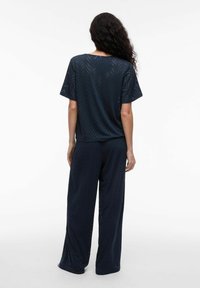 Tums zilo viļņu komplekts, kas sastāv no brīva krekla ar īsām piedurknēm un platiem biksēm, izgatavots no teksturēta auduma ar čevronu rakstu un perforācijām.