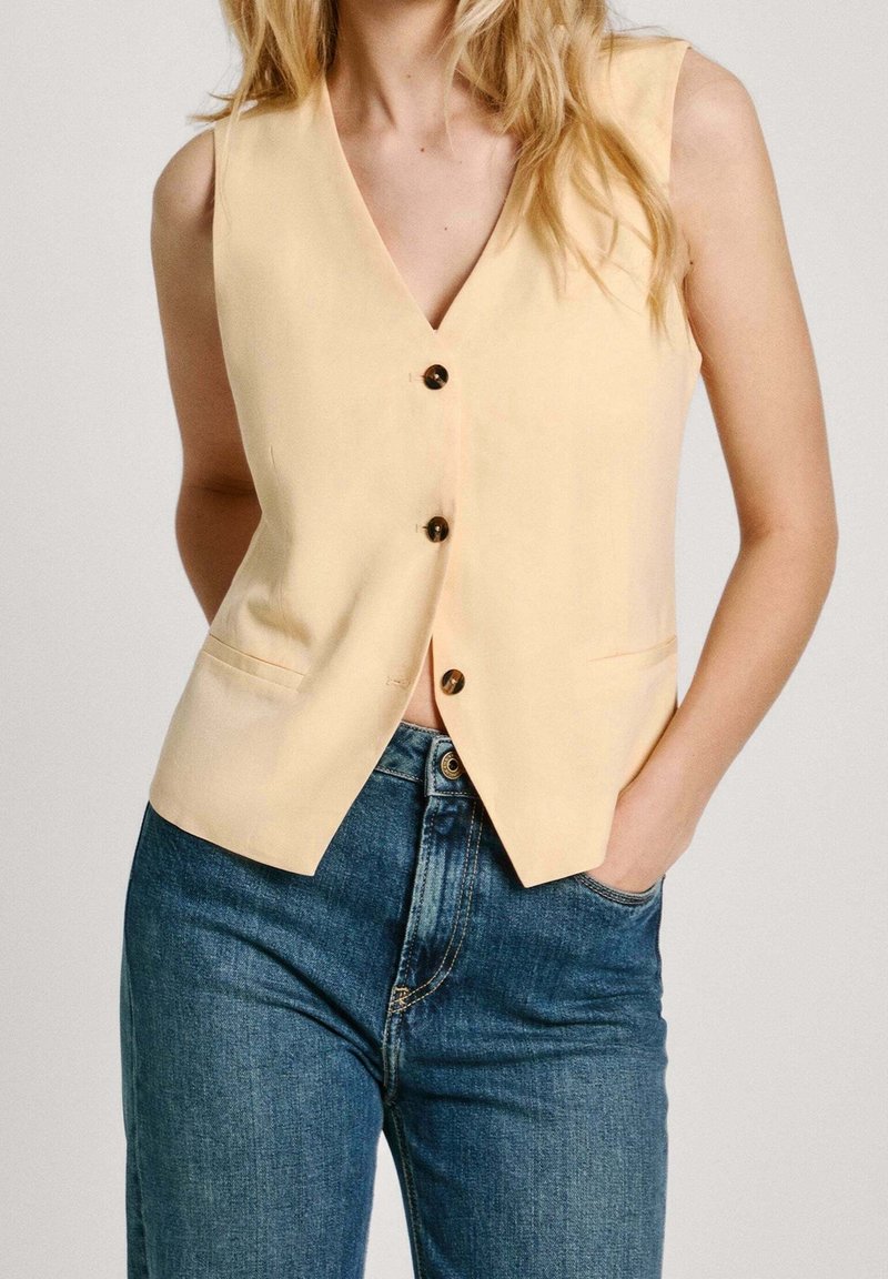Gilet giallo senza maniche con scollo a V profondo, cinque bottoni marroni e due tasche orizzontali, abbinato a jeans in denim blu.