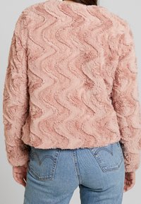 Růžová měkká bunda s vlnitým texturovaným vzorem, dlouhými rukávy a volným střihem, ve spojení se středně modrými džínami.