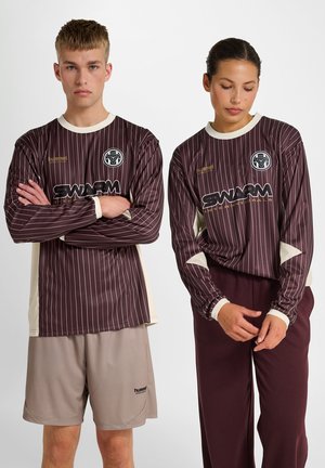 To modeller iført matchende brune langærmede skjorter med smalle striber og "SWARM" logo; den ene i beige shorts, den anden i mørke bukser.