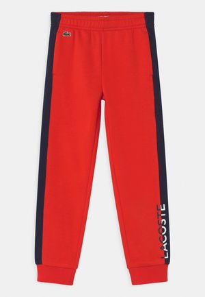 Červené tepláky s elastickým pasem, námořnicky modrými pruhy na stranách a bílým nápisem "LACOSTE" na levé nohavici. Obsahují malé zelené logo.