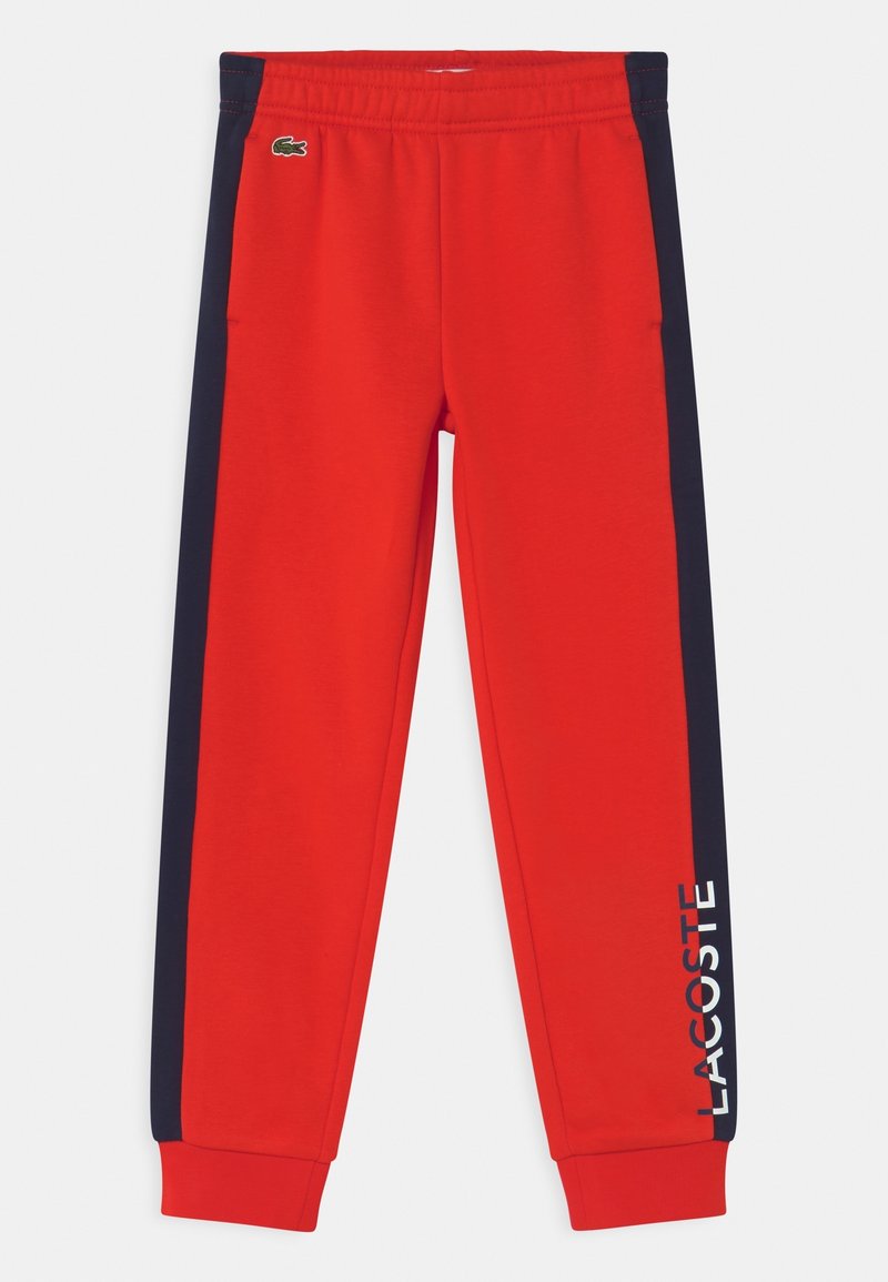 Czerwone spodnie dresowe z elastycznym pasem, granatowymi paskami po bokach i napisem "LACOSTE" w białym kolorze na lewej nogawce. Posiadają małe zielone logo.