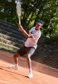 Moški teniški igralec v sivi majici in črnih kratkih hlačah zamahuje s teniškim rekvizitom na glinenem igrišču. Ima obute bele čevlje in nosi temen vizir.