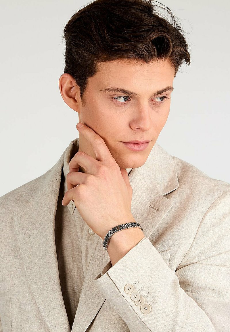 Br Acciaio Bracciale intrecciato nero sul polso di un uomo, con un design testurizzato. Indossa un completo in lino chiaro, che mette in mostra una palette neutra.