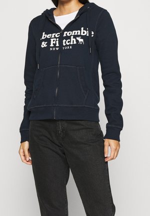 Personne portant un sweat à capuche zippé bleu marine avec le logo Abercrombie & Fitch New York, associé à un jean noir et une chemise blanche en dessous.
