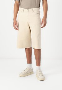 Shorts de mezclilla beige con un corte recto, dobladillo hasta la rodilla, bolsillos frontales y cierre de botón metálico, combinados con zapatillas ligeras.