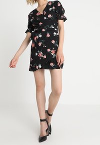 Svart blommig omslagsklänning med korta puffärmar och volangdetaljer. Har rosa och gröna blommönster. Bärs med svarta högklackade skor.