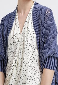 Cárdigan de punto en azul marino, con un punto texturizado y tejido abierto, superpuesto a una blusa crema con lunares negros.