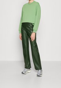 Sudadera verde corta, pantalones de imitación cuero verde oscuro de cintura alta y zapatillas deportivas grises, modelado contra un fondo neutro.