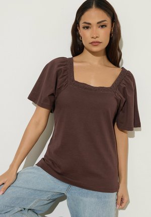 Jeune femme portant un haut marron à manches courtes avec un col carré et un jean bleu clair, debout devant un fond blanc uni.