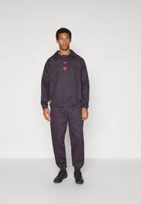 Σκούρο μοβ φόρμα από λεία ύφασμα, που περιλαμβάνει φούτερ με κουκούλα με κόκκινες λεπτομέρειες λογότυπου και ταιριαστά παντελόνια με στενή εφαρμογή. Μαύρα αθλητικά παπούτσια.