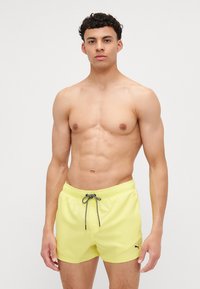 Shorts de baie galbeni cu talie elastică și șnur negru. Prezintă un logo mic pe coapsa stângă. Textură netedă.