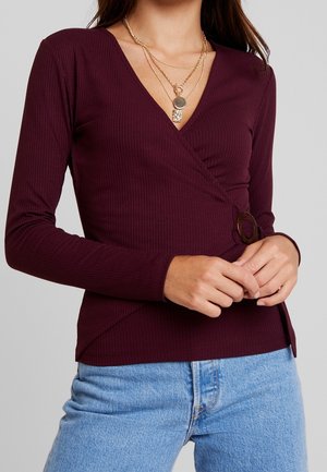 Kvinde iført en rippet, mørk bordeaux omslagsbluse med lange ærmer og spænde, lagdelte guldkæder og lyseblå jeans.