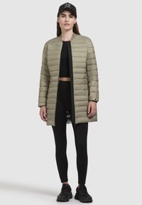Lång, puffer-liknande kappa i ljusgrön med dragkedja framtill. Bärs över en svart crop top och leggings, i kombination med svarta träningsskor och keps.
