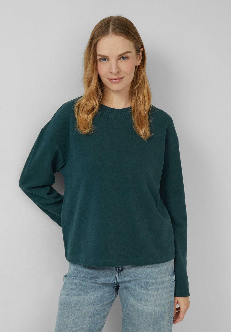 Femme avec de longs cheveux blonds portant un pull vert foncé et un jean bleu clair, se tenant devant un fond gris clair uni.