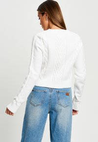 Pull en tricot blanc avec un motif en torsades sur les manches et le dos, poignets et ourlet côtelés, associé à un jean taille haute bleu clair.