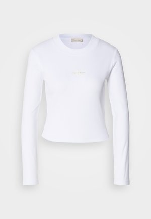 Crop top blanc à manches longues avec un logo Calvin Klein discret au centre, présenté sur un fond gris clair.