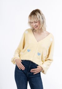 Cardigan giallo fluffy con scollo a V, decorato con applicazioni a forma di cuore nei toni pastello. Ha una chiusura con bottoni e maniche ampie, abbinato a jeans scuri.