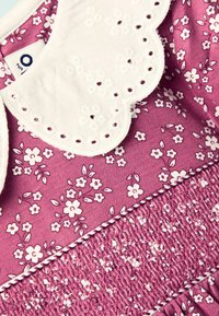 Lila Stoff mit kleinem weißen Blumenmuster, featuring einen gewellten weißen Kragen und Struktursmocking-Details entlang des unteren Randes.