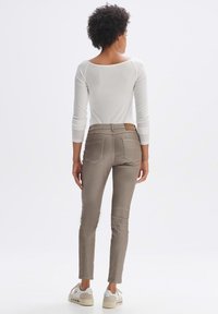 Beige Slim-Fit-Hosen mit Gesäßtaschen, einem Lederpatch am Bund und einer glatten Textur. Kombiniert mit einem weißen Langarmoberteil.