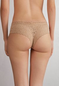 Kantenslips in een huidskleur met een geschulpte tailleband. Het ontwerp heeft ingewikkelde florale patronen en een zachte textuur.