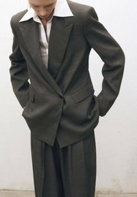Completo su misura grigio scuro con blazer a rever a punta, ampio colletto bianco e pantaloni a gamba larga; texture sottile in tutto il capo.