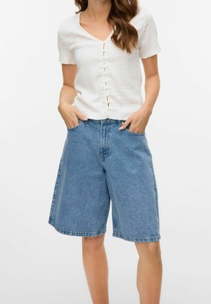 Vrouw draagt een wit, kort mouwtje, met knopen, en loszittende knielange blauwe denimshorts, met handen in de zakken.