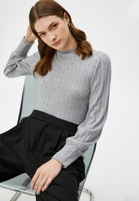 Vrouw met lichtbruin haar, die gouden hoepeloorbellen, een grijs geribbeld langarmtopje en zwart hoge taillebroeken draagt, zittend op een doorzichtige stoel.