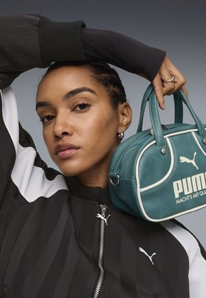 Donna con giacca Puma bianca e nera tiene una borsa Puma turchese vicino al viso su uno sfondo grigio uniforme.