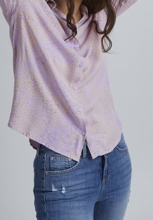 Kvinde iført en lyse-lilla, plettet knap-op bluse og blå denimjeans med let slid på låret.
