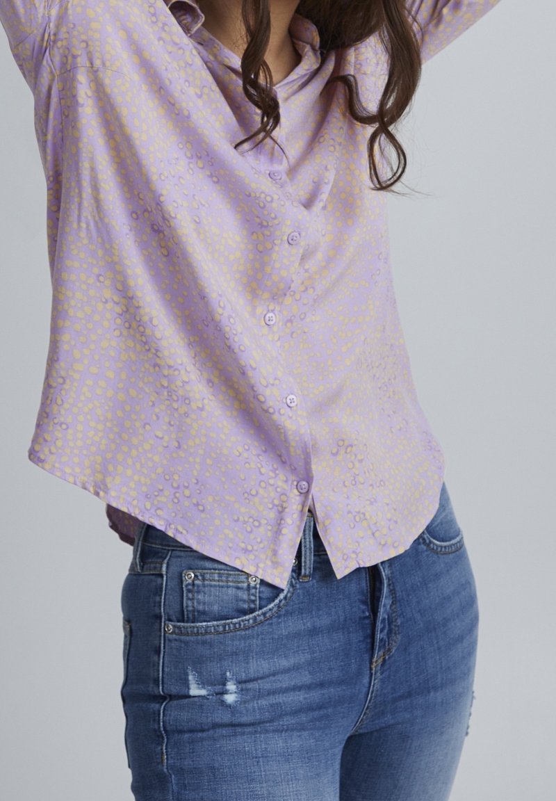 Femme portant un chemisier boutonné violet clair à pois et un jean en denim bleu avec une légère usure au niveau de la cuisse.